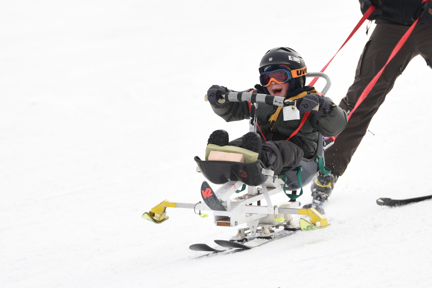 Young participant skiing down the slope in a biski. Participant is bundled up and smiling while holding on to handlebars.
