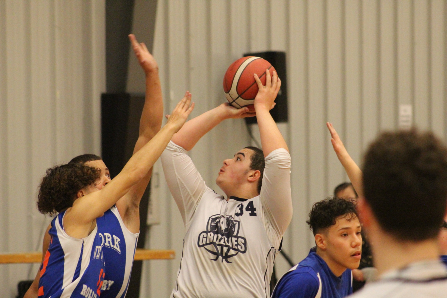 A high school boy in a sports wheelchair holding the ball above his head to shoot as many other players from the opposing team surround him with their hands up.