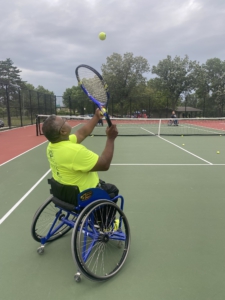 Mike holding his racquet and looking up as he tosses a tennis ball in the air to serve.