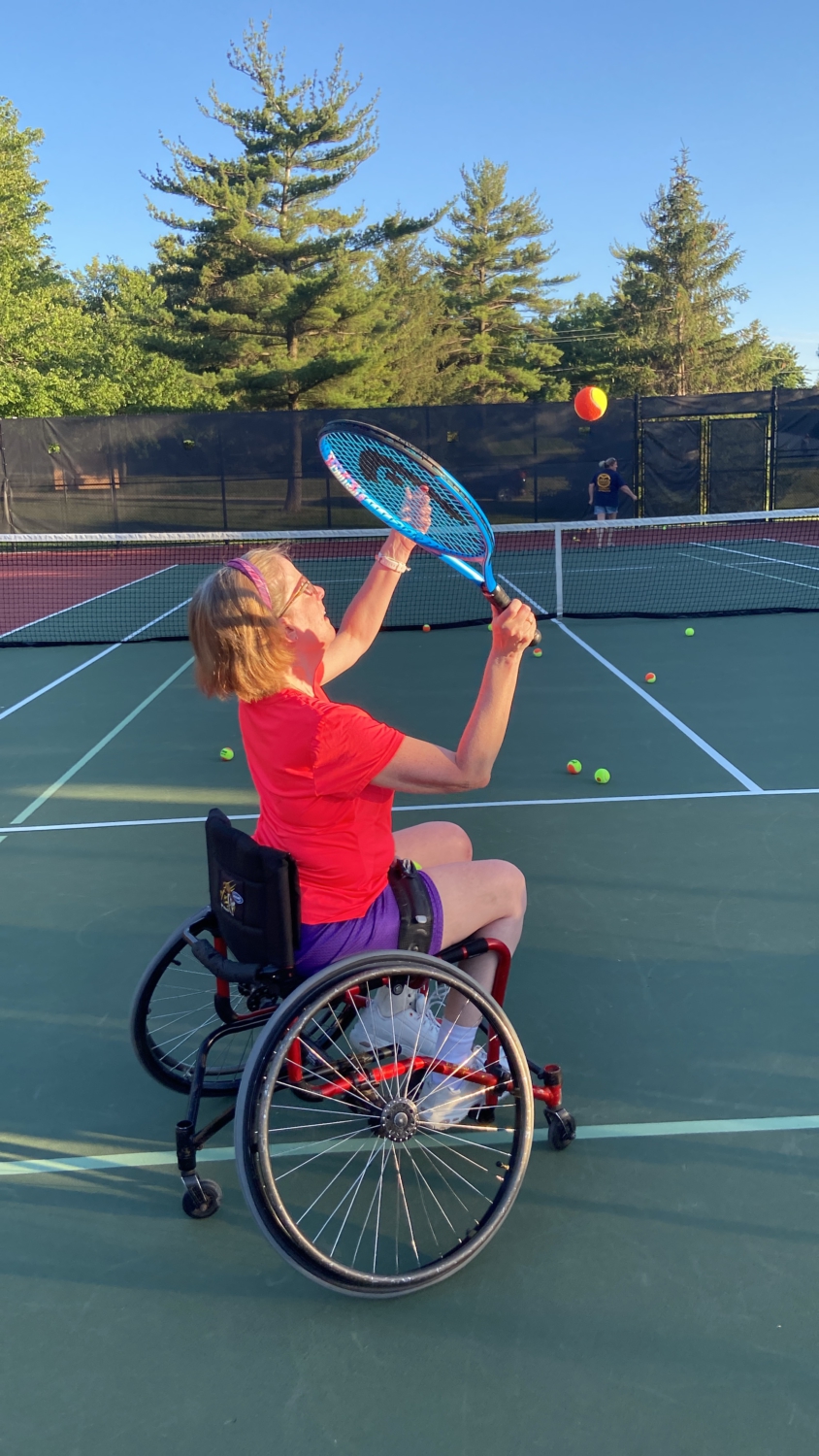 A woman in a sport wheelchair, reaching to serve a tennis ball with her racquet