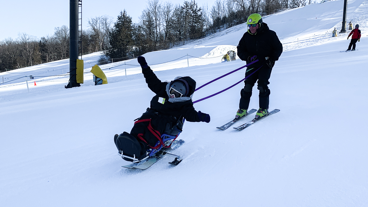 A person all bundled up and sitting in a bi-ski. She is downhill skiing with her hands up in the air. There is an instructor holding a tether behind her and giving turning directions.
