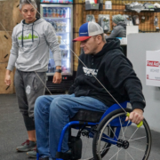 An ASI participant sitting in his everyday chair using the ski machine at the gym at the trainer looks and and gives instructions.