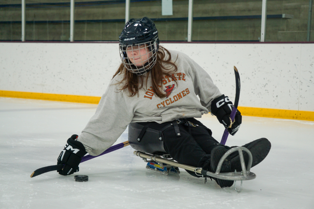 One of the ASI sled hockey participants, Ava, is handling the puck as she turns in her sled. She is wearing a gray Iowa State sweatshirt, black pants, a helmet, and gloves.