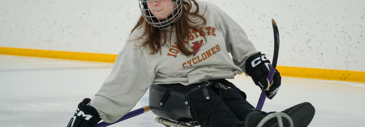 One of the ASI sled hockey participants, Ava, is handling the puck as she turns in her sled. She is wearing a gray Iowa State sweatshirt, black pants, a helmet, and gloves.