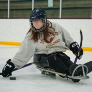 One of the ASI sled hockey participants, Ava, is handling the puck as she turns in her sled. She is wearing a gray Iowa State sweatshirt, black pants, a helmet, and gloves.