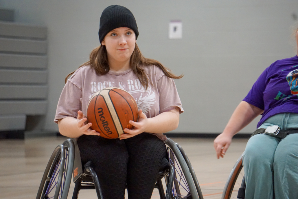 Ava is holding the basketball in her sports chair with a grin on her face.