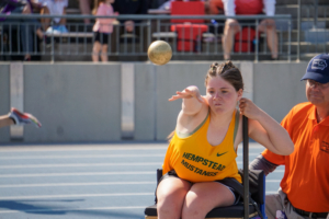 Ally sitting in a shot put chair, throwing the implement in her yellow team track jersey.