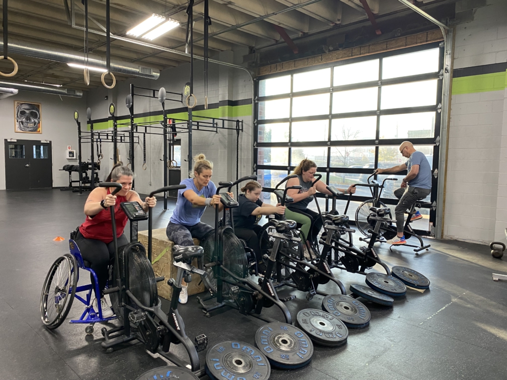 Four women working out next to each other. They are all seated and using a stationary bike with their arms.