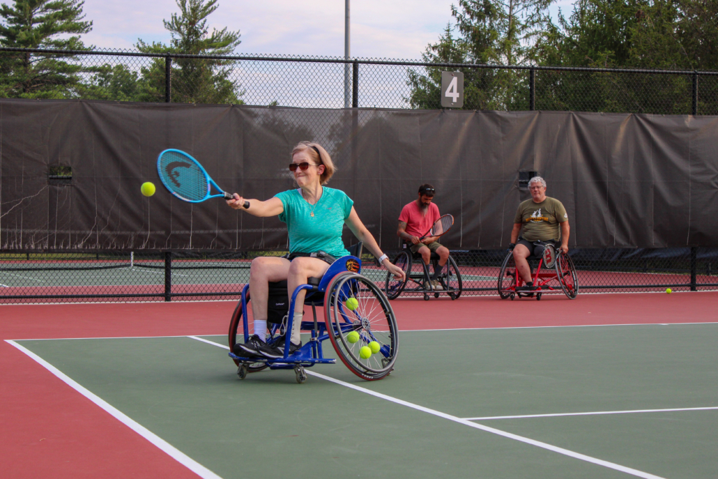 Woman in a sports wheelchair swinging a tennis racquet towards the ball in the back right corner of the court.